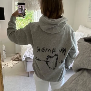 Grå hoodie med tryck 'Mi Amor' - Säljer en grå hoodie med svart tryck på ryggen där det står 'Mi Amor' och ett hjärta med pil. Hoodien har huva med snörning och en stor magficka. Perfekt för chill dagar och enkel att styla med jeans eller mjukisbyxor.