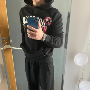 Svart hoodie från Hoodrich med tryck - Svart hoodie från Hoodrich med vit och röd text och logga framtill. Stor ficka på magen och huva med dragsko. Stora tryck och patchar ger en streetwear-känsla. Perfekt för dig som gillar en sportig och avslappnad stil. Andvänd ett par gånger men fortfarande i topp skick (9/10). Storlek S. Nypris 899