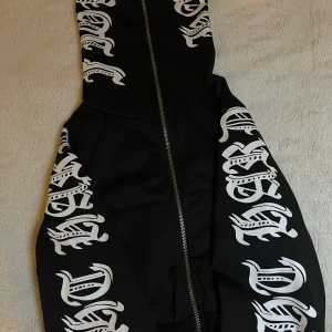 Svart zip up hoodie  - Svart zip up hoodie med stora vita tryck i gotisk stil längs hela luvan och ärmarna. Koftan är en zip up hoodie och passar alla olika outfits. Pris kan diskuteras🤍