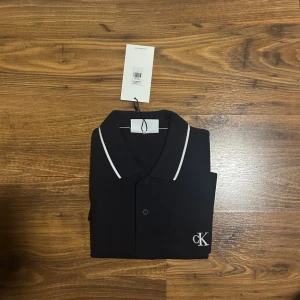 Svart pikétröja från Calvin Klein - Snygg svart pikétröja från Calvin Klein med vit kant på kragen och ärmsluten. Tröjan har korta ärmar och klassisk pikékrage. Diskret CK-logga broderad på bröstet. Perfekt för en stilren och enkel look.