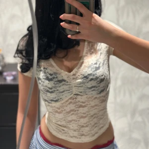 Vit spets croppad topp - Säljer en vit croppad topp i spets med v-ringning och korta ärmar. Toppen är i transparent spetsmaterial och har en figurnära passform. På bilden använder jag en säkerhetsnål för att uppnå tightare passform.