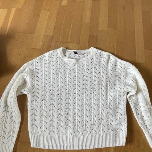 Vit stickad tröja från H&M Divided - Säljer en vit stickad tröja från H&M Divided med hålmönster och rund hals. Tröjan har långa ärmar och ribbade muddar vid ärmslut och nederkant. Perfekt för lager-på-lager och passar till många olika stilar.