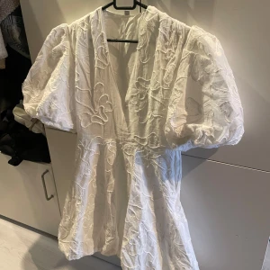 Vit kortklänning med puffärmar och broderade blommor - Superfin vit kortklänning med djup v-ringning, puffiga kortärmar och broderade blommor. Klänningen har en luftig passform och är perfekt för sommarens tillfällen. Materialet känns lätt och har en vacker struktur.