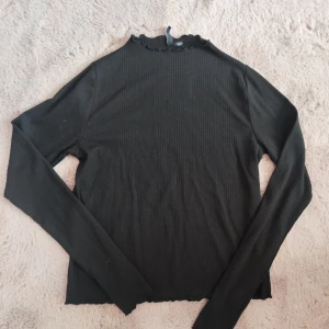 Svart långärmad topp med volangkant - Säljer en svart långärmad topp med volangkant vid hals och ärmslut. Toppen har en tight passform och är perfekt att styla till olika outfits. Enkel och stilren design som passar till mycket.