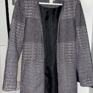 Svartvit mönstrad kappa från H&M - Säljer en svartvit kappa från H&M med grafiskt mönster. Jackan har lång ärm, rak passform och är helfodrad med svart innerfoder. Perfekt för dig som gillar stilrena och tidlösa plagg.