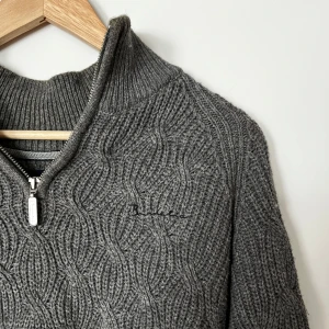 Grå stickad tröja med dragkedja från Bondelid - Mysig grå stickad tröja från Bondelid med halv dragkedja och diskret mönster. Tröjan har hög krage och långa ärmar, perfekt för kyliga dagar. Klassisk och stilren design som passar till mycket.