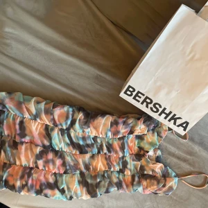 Färgglad omlottklänning från Bershka - Säljer en kort, ärmlös klänning från Bershka med färgglatt mönster i pastelltoner. Klänningen har omlottdetalj och tunna axelband, perfekt för varma dagar.