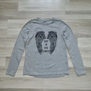 Grå långärmad tröja med tryck från Ellos - Säljer en grå långärmad tröja från Ellos med svart tryck av vingar och texten 'Sky is the limit' på framsidan. Tröjan är i mjukt bomullsmaterial och har rund hals. Perfekt för en avslappnad stil.
