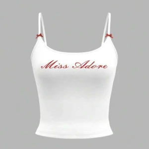 Vit croppad linnetopp med text - Säljer en vit croppad linnetopp med tunna axelband och röda rosetter. Toppen har texten 'Miss Adore' i rött framtill och är perfekt för sommaren.