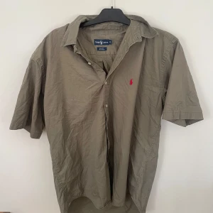 Beige kortärmad skjorta från Ralph Lauren - Säljer en klassisk beige kortärmad skjorta från Ralph Lauren i 100% bomull. Skjortan har knappar framtill och den ikoniska broderade loggan i rött på bröstet. Perfekt för en avslappnad och stilren look.