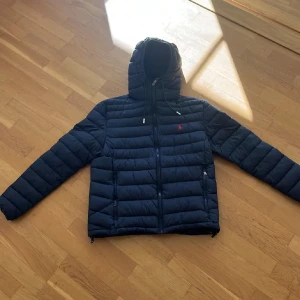 Mörkblå dunjacka från Polo Ralph Lauren - Säljer en mörkblå dunjacka från Polo Ralph Lauren med huva och dragkedja. Jackan har ett rött broderat logomärke på bröstet och två sidofickor med dragkedja. Perfekt för kyliga dagar.