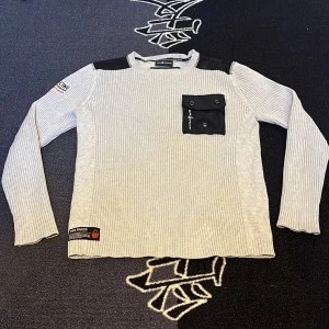 Ljusgrå ribbad tröja från Sail Racing - Ljusgrå långärmad tröja från Sail Racing med ribbad struktur och svarta detaljer på axlar och bröstficka. Bröstfickan har knappstängning och logga. 