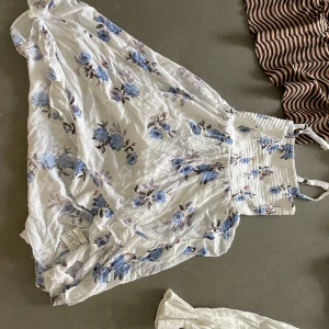 Vit blommig klänning med smock i midjan - Supersöt vit klänning med blått blommönster och bred smock i midjan. Klänningen är luftig och har en lätt, flowy känsla – perfekt för varma dagar. 