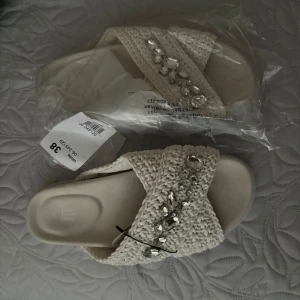 Vita sandaler med pärldetaljer - Säljer ett par vita sandaler från dinsko med breda, stickade band och dekorativa pärlor och stenar på ovansidan. Skorna har platt sula och öppen tå, perfekta för varmare dagar.