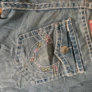 Blå true religion jeans med färgglada diamanter och kontrastsömmar - Säljer ett par blå jeans med vita kontrastsömmar och dekorativa nitar i rosa, gult och blått på bakfickan och framtill. Jeansen har klassisk femficksdesign och är bootcut. 