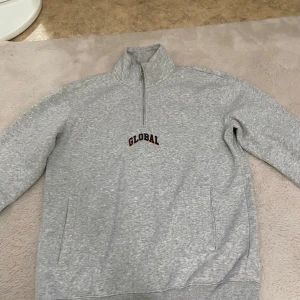 Grå halvzip sweatshirt från H&M - Gråmelerad sweatshirt från H&M med halv dragkedja och hög krage. Broderad text 'GLOBAL' på bröstet. Tröjan har långa ärmar och två fickor framtill. Perfekt för en avslappnad stil.