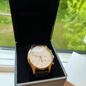 Emporio Armani herrklocka med svart läderband - Stilren klocka från Emporio Armani med vit urtavla, guldfärgad boett och svart läderarmband. Klockan har tre visare, datumvisning och klassisk design. Levereras i originalask med Emporio Armani-logga. Klocka funkar för tillfället inte men behöver bara byta batteri. 