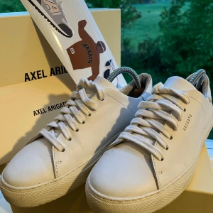 Vita sneakers från Axel Arigato - Säljer ett par stilrena vita sneakers från Axel Arigato med klassisk design. Skorna har rund tå, platt sula och snörning framtill. Tillverkade i skinn med diskret logga på sidan. Perfekta för en clean och modern look.