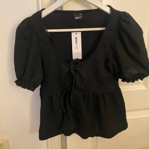 Svart blus med puffärm från Gina Tricot - Säljer en svart blus från Gina Tricot med puffiga korta ärmar och knyt framtill. Aldrig använd och har prislapp kvar🩷