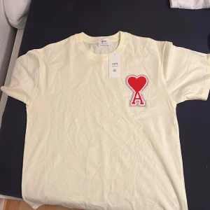 Vit t-shirt från Ami med rött hjärta - Säljer en vit t-shirt från Ami Alexandre Mattiussi med ett stort rött hjärta och bokstaven A broderat på bröstet. Klassisk rund hals och normal passform. Perfekt för dig som gillar stilrena plagg med en twist.