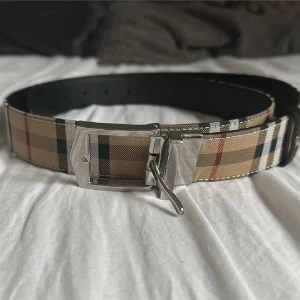 Rutmönstrat bälte från Burberry - Säljer ett klassiskt bälte från Burberry med det ikoniska rutmönstret i beige, svart, vitt och rött. Bältet har ett silvrigt spänne och är tillverkat i skinn. Perfekt accessoar för att lyfta din outfit.