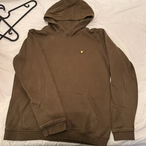 Brun hoodie från Lyle & Scott Junior - Säljer en brun hoodie från Lyle & Scott Junior med klassisk huva och magficka. Tröjan har ett broderat gult logomärke på bröstet och ribbade muddar vid ärmslut och nederkant. Perfekt för en avslappnad stil.
