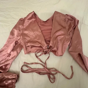 Rosa omlottopp med knytband - Trendig rosa omlottopp i glansigt material med långa puffärmar och knytband i midjan. Perfekt för att skapa en snygg och feminin look. Toppen har en croppad passform och v-ringad hals.