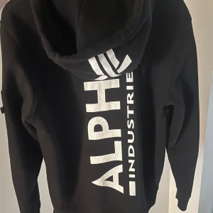 Svart hoodie från Alpha Industries - Säljer en svart hoodie från Alpha Industries med stor vit logga på ryggen och mindre logga på bröstet. Tröjan har huva med dragsko, magficka och en ärmficka med dragkedja och röd tag. Perfekt för en cool och avslappnad stil. Pris kan diskuteras vid snabb handel.