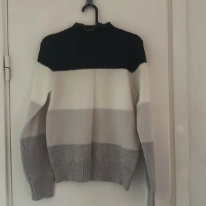 Randig stickad tröja i svart, vitt och grått - Säljer en stickad tröja med breda ränder i svart, vitt och olika nyanser av grått. Tröjan har lång ärm och ribbade muddar vid ärmslut och nederkant. Perfekt för kyligare dagar.
