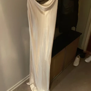 Vit långklänning med öppen rygg och tunna band - Säljer en elegant vit långklänning med draperad halsringning och öppen rygg med tunna korsade band. Klänningen har en slät, glansig yta och faller snyggt längs kroppen. Perfekt för speciella tillfällen.