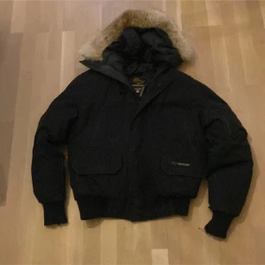 Svart vinterjacka med päls från Canada Goose - Säljer en svart vinterjacka från Canada Goose med huva och pälsdetalj. Jackan har två stora fickor framtill, ribbade muddar och klassisk Canada Goose-logga på ärmen. Perfekt för kalla dagar. Jackan har två små skrapor vid slutändan av armarna men forfarande en fin vinterjacka!