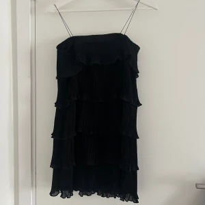 Svart volangklänning med tunna axelband - Säljer en svart klänning med tunna axelband och flera lager volanger. Klänningen är axelbandslös och har en luftig, plisserad design som ger en cool och trendig look. Perfekt för dig som gillar stilrena och eleganta plagg.