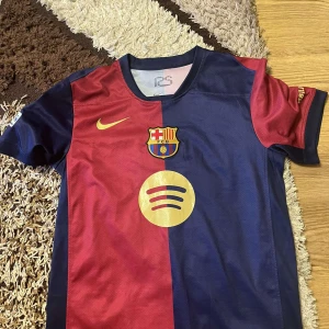 FC Barcelona matchtröja Lamine Yamal 19 - Säljer en officiell FC Barcelona matchtröja med Lamine Yamal och nummer 19 på ryggen. Tröjan är röd och blå med guldiga detaljer, klubbmärke och sponsorer. Kortärmad modell i lätt och ventilerande material, perfekt för fotboll eller samling.