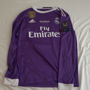 Lila Real Madrid långärmad fotbollströja Ronaldo 7 - Säljer en långärmad lila Real Madrid fotbollströja från Adidas med vita detaljer. Tryck med Ronaldo 7 på ryggen och klubbmärke på bröstet. Champions League-märke på ärmen och Fly Emirates sponsortryck framtill.
