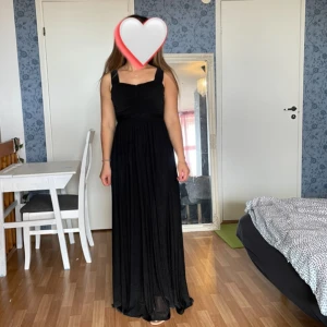 Svart långklänning med breda axelband - Säljer en elegant svart långklänning med breda axelband och lätt rynkad överdel. Klänningen har en klassisk och stilren design som passar perfekt till fest eller bal. Materialet är mjukt och faller snyggt längs kroppen.