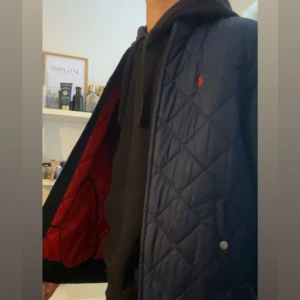Blå quiltad väst från Polo Ralph Lauren - Säljer en mörkblå quiltad väst från Polo Ralph Lauren med röd insida och diskret logga på bröstet. Västen har dragkedja och tryckknappar framtill samt ribbade detaljer vid midjan. Perfekt för lager-på-lager-stil.