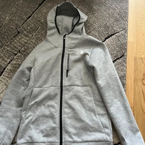 Grå hoodiejacka från The North Face XS/S - Säljer en grå hoodiejacka från The North Face med dragkedja och huva. Jackan har en bröstficka med dragkedja och två stora fickor framtill. Perfekt för kyliga dagar och enkel att matcha med det mesta.