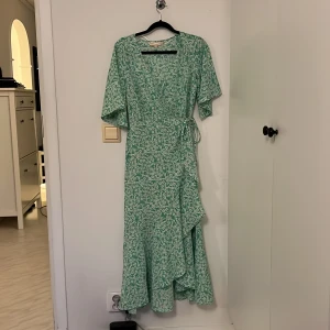 Grön blommig omlottklänning från Pernilla Wahlgren - Superfin grön omlottklänning från Pernilla Wahlgren med vitt blommönster. Klänningen har volangdetaljer, knyt i midjan och korta ärmar. Perfekt för vår och sommar, och har en liten guldig knappdetalj.