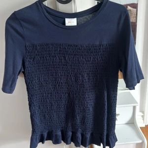 Mörkblå topp med smock och volangkant - En mörkblå topp med smockad framsida och baksida samt volangkant nedtill. Toppen har rund halsringning och halvlånga ärmar. Perfekt för en stilren och trendig look.