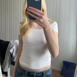 Vit ribbad topp med spetskant - Säljer en söt vit ribbad topp med korta ärmar och fin spetskant längs urringningen. Toppen har en figurnära passform och är perfekt till sommaren. Materialet är mjukt och stretchigt för en bekväm känsla.