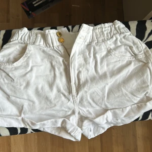 Vita shorts från H&M med guldknappar - Snygga vita shorts från H&M med två guldiga knappar framtill och dragkedja. Shortsen har resår i midjan, två fickor fram och två bak. Perfekta för varma dagar och enkel att matcha med olika toppar. Inte använd någon gång. 