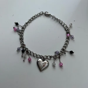Silverfärgat armband med hjärthänge och pärlor - Charmigt armband i silverfärgad metall med ett hjärthänge i mitten. Armbandet är dekorerat med små pärlor i rosa, lila, svart och vitt som hänger längs kedjan. Perfekt accessoar för att ge din stil en söt och lekfull touch.
