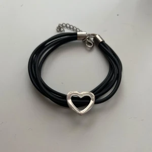 Svart armband med silverfärgat hjärta - Snyggt armband med flera svarta band och ett silverfärgat hjärta i mitten. Justerbart spänne i metall. Perfekt accessoar för att ge din outfit en stilren touch.