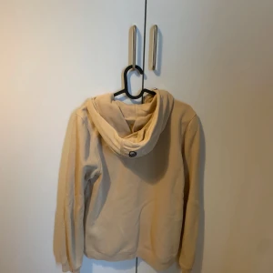 Beige Hoodie från Sail Racing - Beige hoodie med huva från Sailracing. Hoodien har dragkedja framtill och en bröstficka med dragkedja. Perfekt för lager-på-lager och svalare dagar. Mjuk och skön känsla.