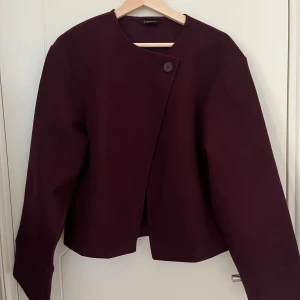 Vinröd kort kappa med asymmetrisk knäppning - Kort vinröd kappa med rund halsringning och en knapp framtill för asymmetrisk stängning. Använd 1 gång i höstas, så den är som gott som i nyskick.