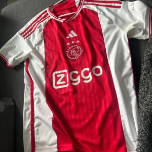 Ajax Amsterdam fotbollströja - Ajax hemmatröja från Adidas med klassiskt rött och vitt färgschema. Tröjan har korta ärmar, Ajax klubbmärke och sponsortryck på bröstet samt tre stjärnor ovanför loggan. Ikoniska Adidas-ränder på axlarna.