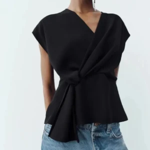 Svart omlottopp från Zara - Snygg svart omlottopp från Zara med knyt i midjan och v-ringad hals. Toppen har korta ärmar och en elegant, stilren design som passar perfekt till jeans eller kjol.