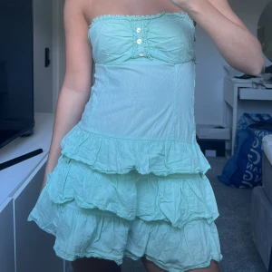 volangklänning  - Supersöt ljusgrön axelbandslös klänning med volanger och dekorativa knappar framtill. Klänningen har en kort längd och smockad rygg för extra komfort. Perfekt för varma dagar och somriga tillfällen!