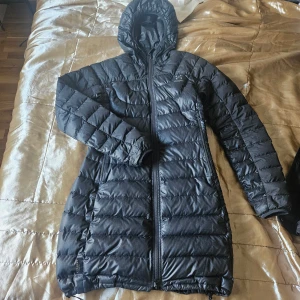 Svart lång dunjacka från Haglöfs - Säljer en svart lång dunjacka från Haglöfs med huva och dragkedja framtill. Jackan är tillverkad i Pertex Quantum-material som är lätt och slitstarkt. Perfekt för kyliga dagar och har en klassisk, stilren look.