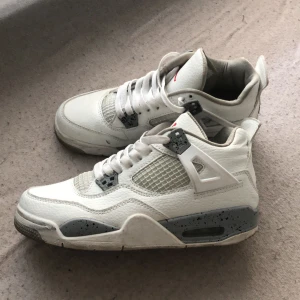 Vita Nike Air Jordans 4 Retro - Säljer ett par klassiska Nike Air Jordans 4 Retro i vitt med grå och svarta detaljer samt röd Jumpman-logga på plösen. Skorna har nätpaneler på sidorna, snörning och en robust sula med synlig luftdämpning.